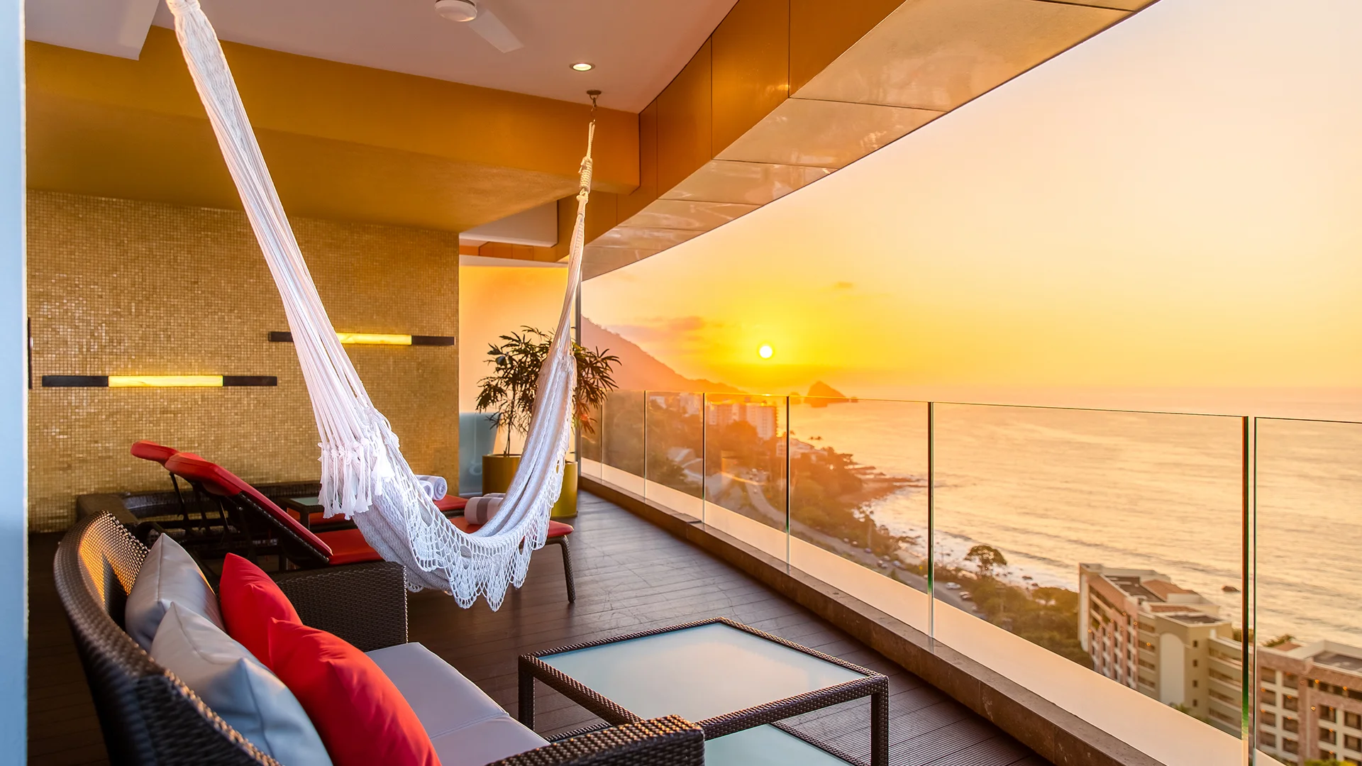 Sunset terrace at Hotel Mousai Puerto Vallarta, an adults-only luxury MICE venue in Mexico for meetings, incentives, conventions, and exhibitions, featuring a white hammock, wicker lounge chairs with red and gray pillows, coffee table, glass railing, and breathtaking views of the Pacific Ocean, coastline, and Sierra Madre mountains ideal for unwinding during incentive travel programs.